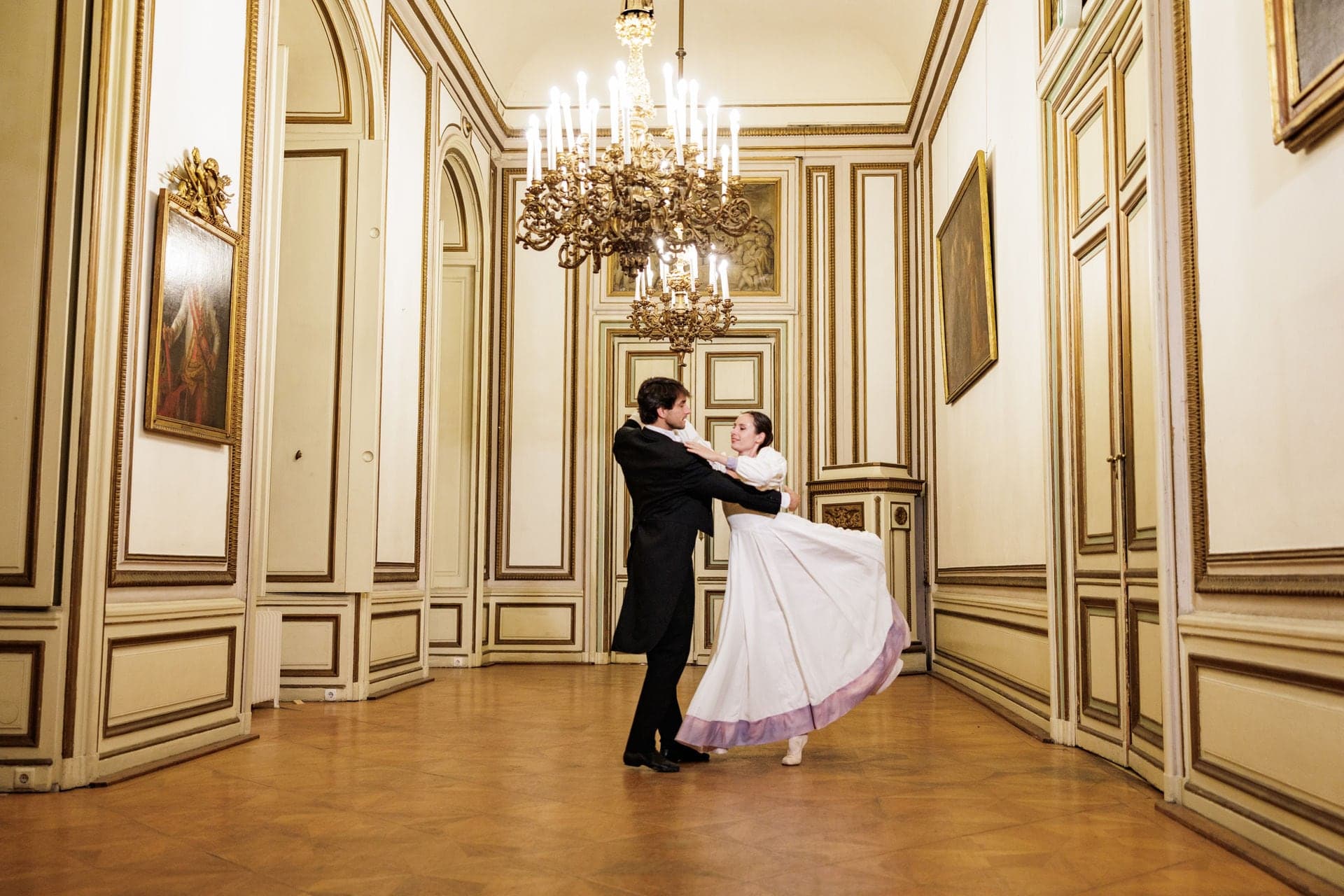 Waltzing couple in tailcoat and white ball gown in a chandeliered salon
